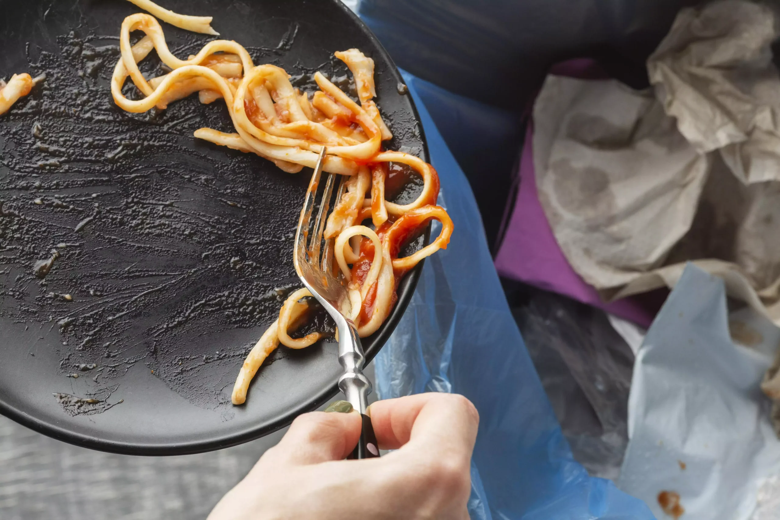 Main de personne tenant une fourchette pour jeter des spaghettis à la sauce tomate d'une assiette noire dans une poubelle bleue, illustrant les pertes alimentaires.