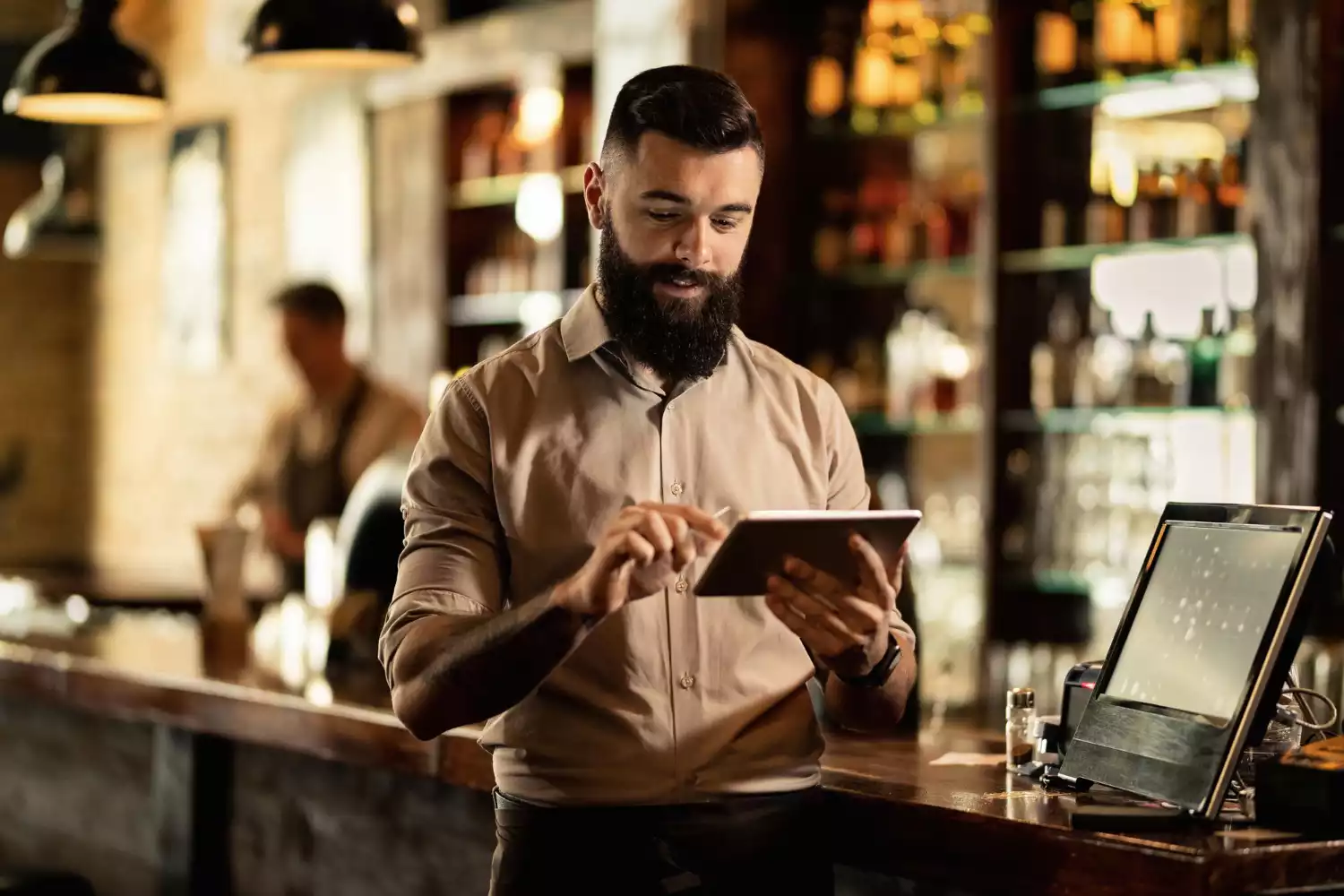 Serveur barman barbu utilisant une tablette tactile connectée au Module Service de Gusto pour prendre une commande près du comptoir d'un restaurant.